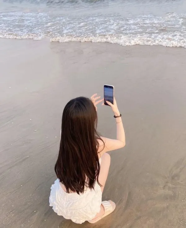 anh-gai-di-bien-che-mat-12 Nàng ngồi sát nước, cầm điện thoại chụp ảnh, tóc thả lưng, vẻ kín đáo dễ thương.