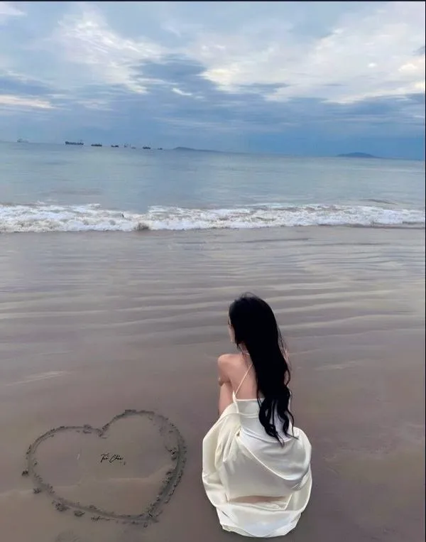 anh-gai-di-bien-che-mat-5 Cô gái ngồi trên cát trắng vẽ trái tim, tóc phủ lưng, ánh nhìn giấu kín sau vai.